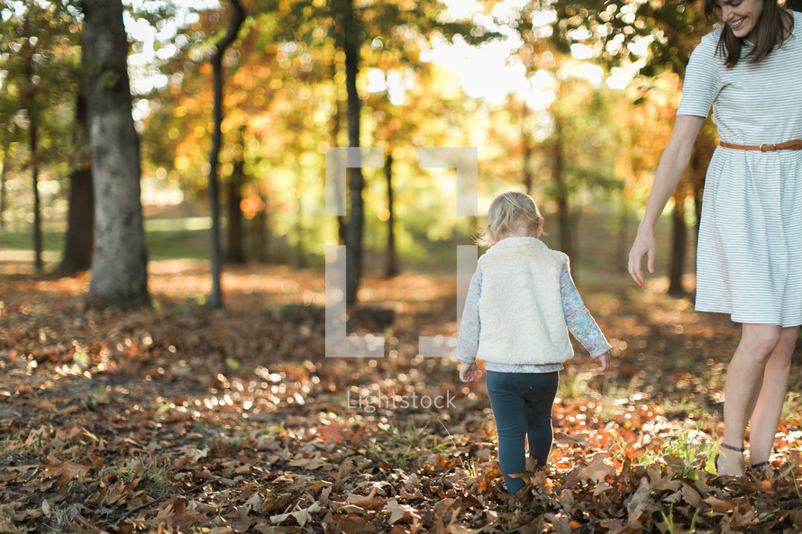 a mother walking with her toddler daughter through fall leaves 