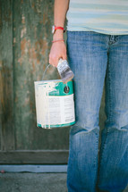Person holding a old paint can and paint brush standing in front of an old green wooden wall