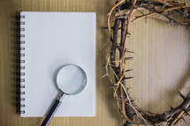 blank notepad, magnifying glass, and crown of thorns on a wood background 