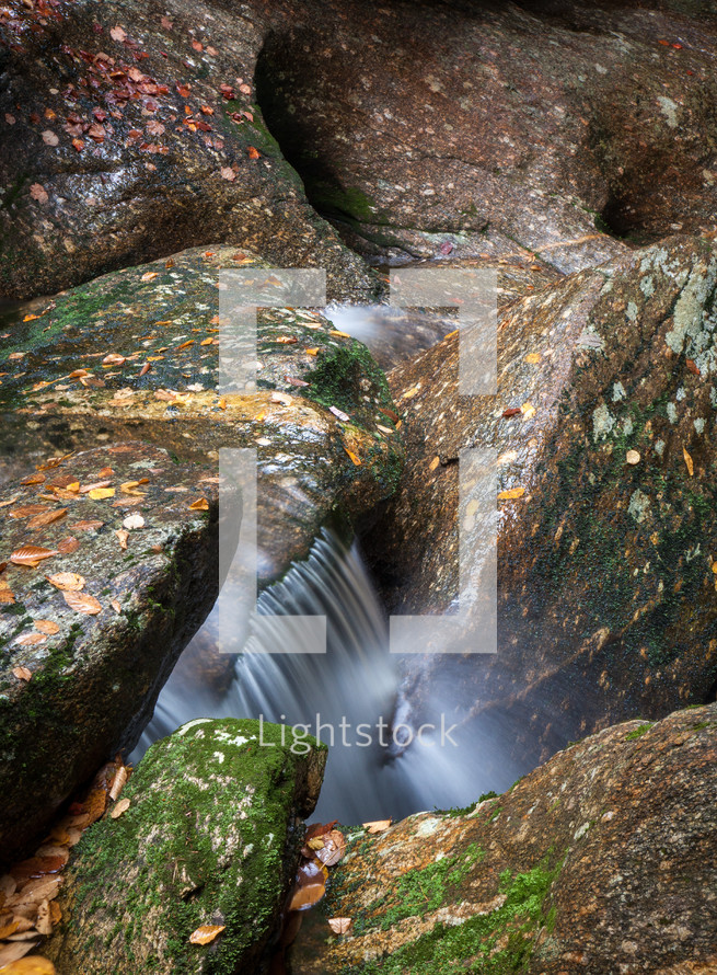 Silky waterfall cascading down large rocks with fallen autumn leaves and moss on them