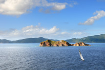 Rock islands in a body of water, a sailboat in the water and mountains in the background