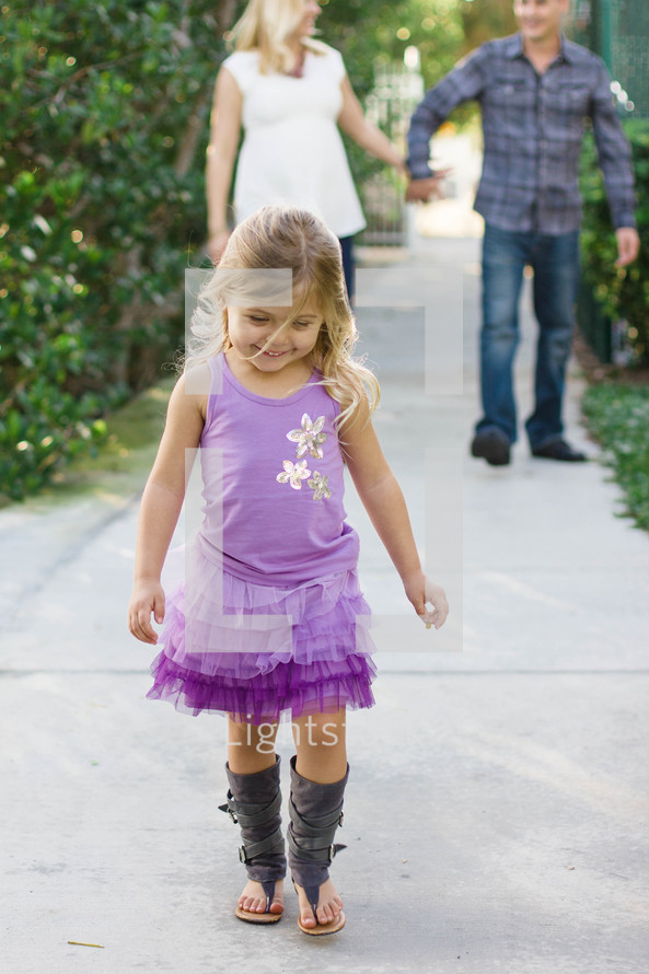 A little girl walking along the sidewalk in front of her parents.