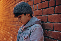a boy leaning against a brick wall looking down 