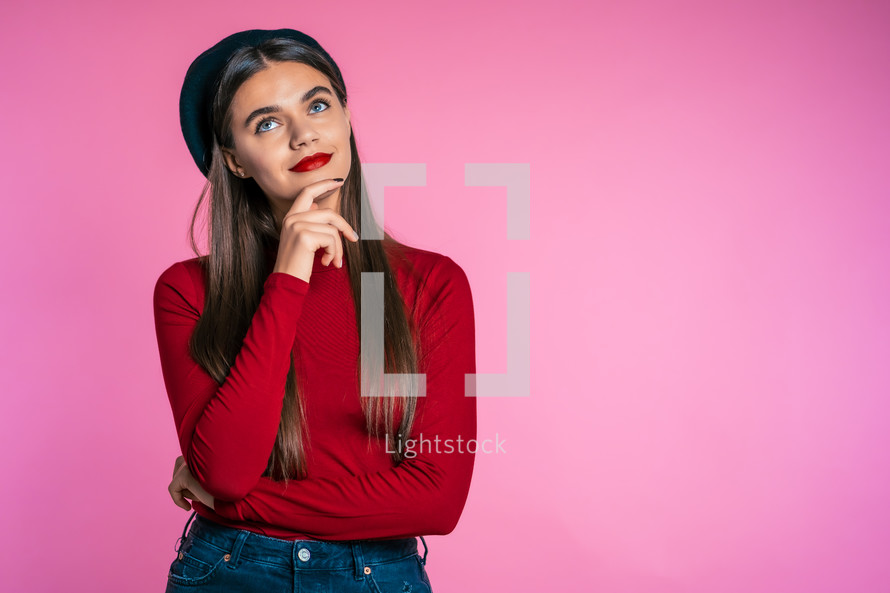 Serious girl in red wear. Portrait of young thinking pondering pretty woman on pink background.
