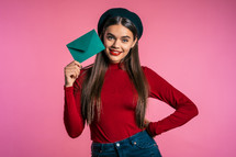 Portrait of young beautiful woman in red wear and hat with green envelope on pink studio background
