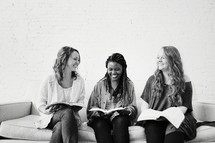 young women sitting on a couch talking, laughing, and reading bibles 