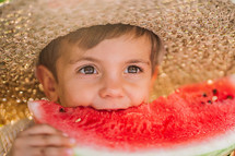 Cute little boy eating slice of juicy watermelon sitting on natural green garden background. Adorable kid in straw hat enjoying summer fresh melon fruit with smile. High quality photo