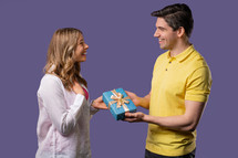 Young american couple on violet studio background. White man gives gift with bow to wife or girlfriend. Birthday, celebration, surprise, anniversary concept. High quality