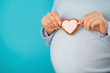 Pregnant woman holding heart-shaped cookie on tummy belly background. Young girl in blue wearing expecting baby. Maternity, motherhood, pregnancy, love concept