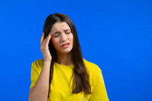 Portrait of woman having headache, blue studio portrait. Girl in yellow t-shirt putting hand on head. Concept of migraine problems, medicine, illness. High quality photo