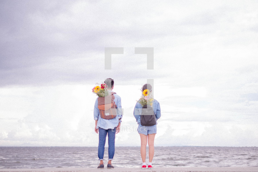 couple standing on a shore with flowers in their backpacks 
