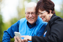 elderly couple sitting on a park bench looking at a cellphone screen 