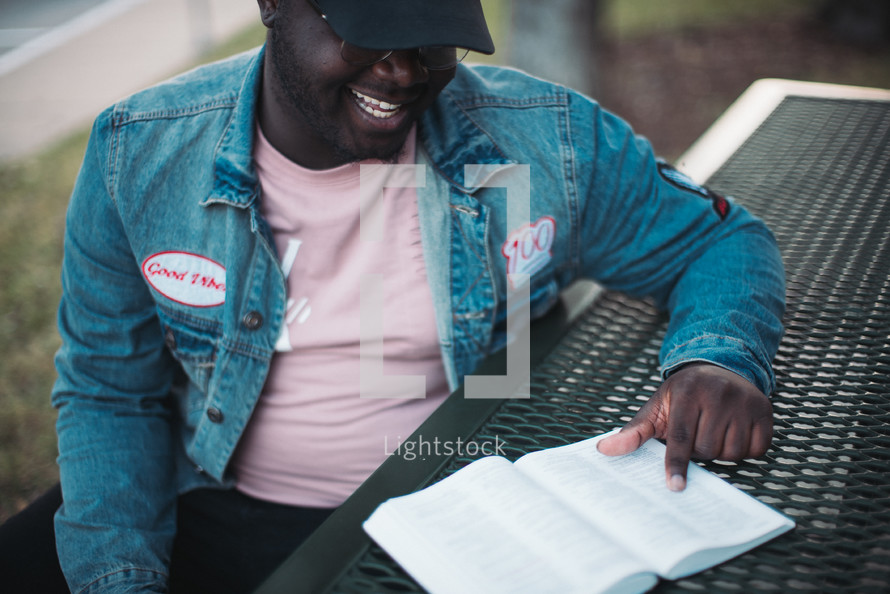 a man sitting down at a table outdoors reading a Bible 