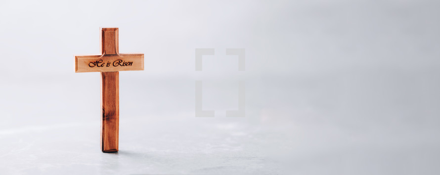 Wooden cross with text "He is risen" on grey background.