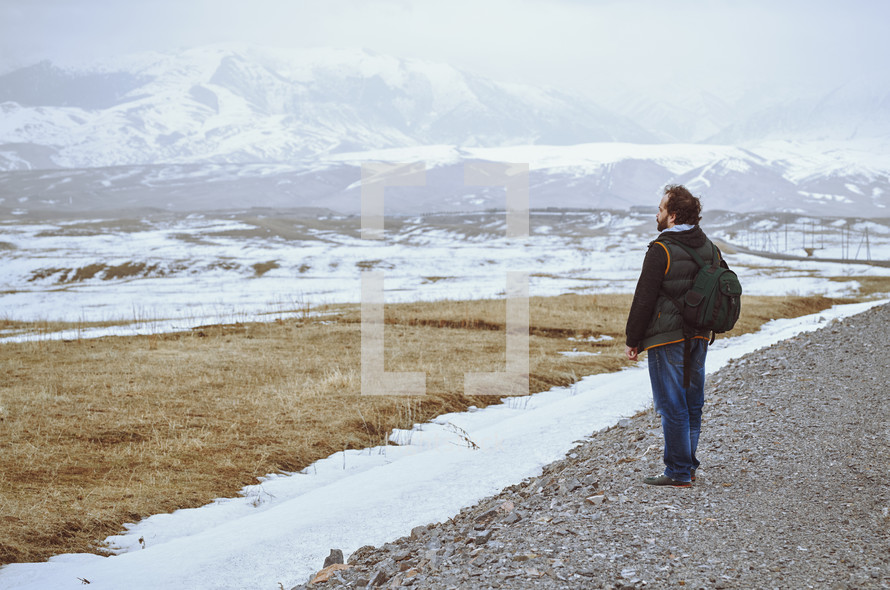 Tourist wearing backpack and standing at the rural road while looking at the winter mountains