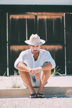 a man sitting on the curb wearing a cowboy hat and flip flops 
