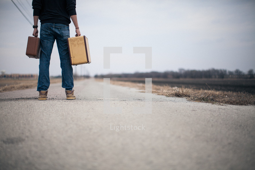 man holding suitcases looking down a long road
