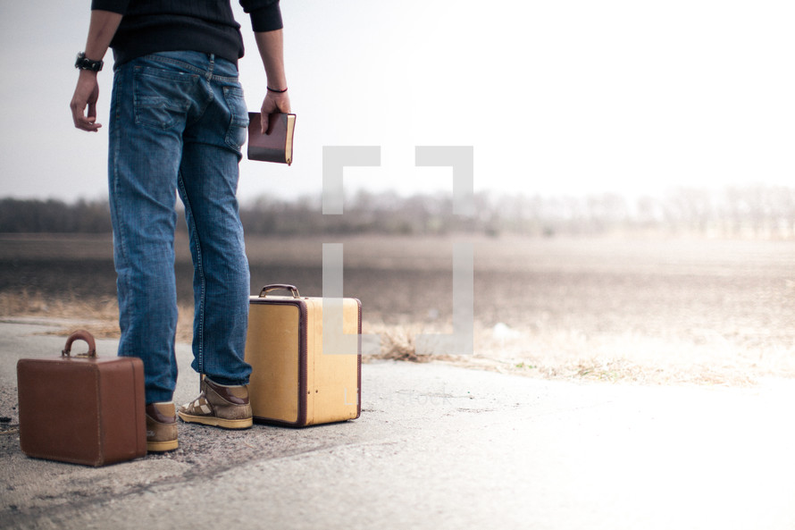man standing next to a suitcase holding a Bible and looking down a road wondering where to go from here
