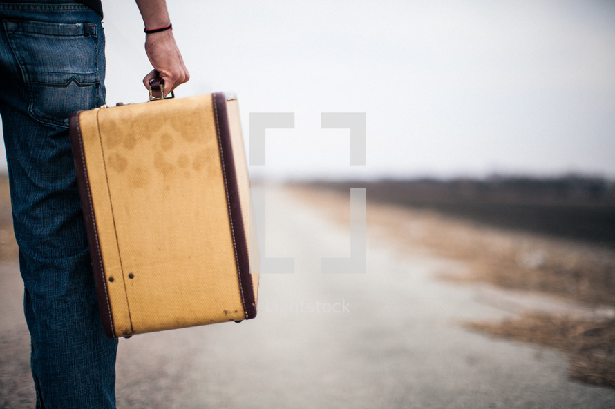 close-up of a man carrying a suitcase looking down a long road
