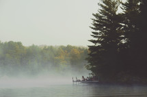 Morning mist over a lake with large pine trees. Dock and Muskoka chairs.