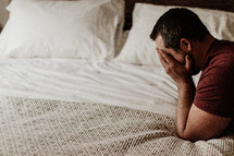 a man covering his face kneeling in prayer over a bed 