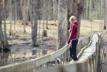 A young woman alone on a pedestrian bridge in a wooded area.