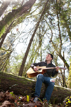 man sitting on a fallen tree in a forest playing his guitar