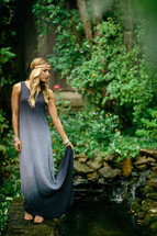 a young blonde woman with a headband and long dress standing next to a koi pond 