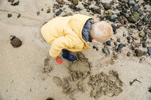 a toddler boy playing in sand on a beach in a coat 