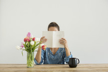 a woman sitting at a desk with a mug of coffee and a vase of tulips reading a Bible 