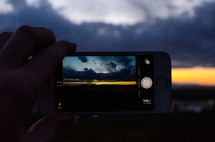 man taking a picture of a cloudy sky at dusk with his cellphone 