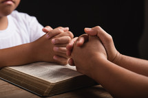 A child and mother praying with hands folded over a Bible