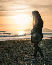 a woman in a coat standing on a rocky beach at sunset 