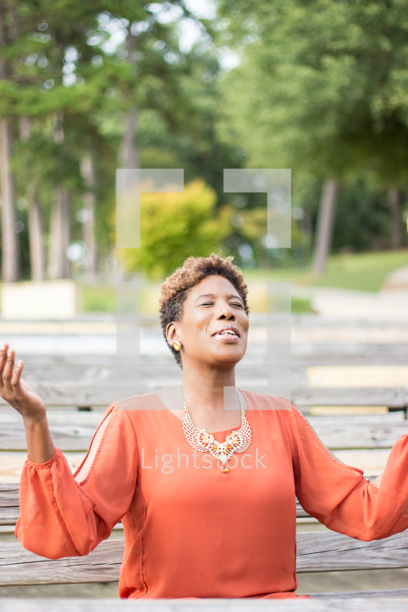 a woman with raised hands praising God sitting on a park bench 