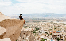 a man sitting at the edge of a cliff looking out at the western town below 