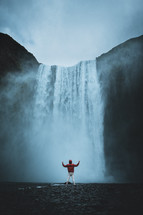person with raised hands standing in front of a waterfall 