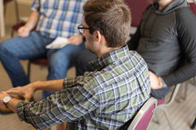 Young adult men sitting in circle during Bible study at church, discussing sermon, dark haired man in green plaid shirt in focus