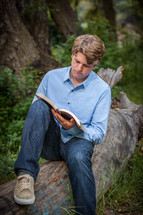 man reading a book sitting on a fallen tree outdoors 