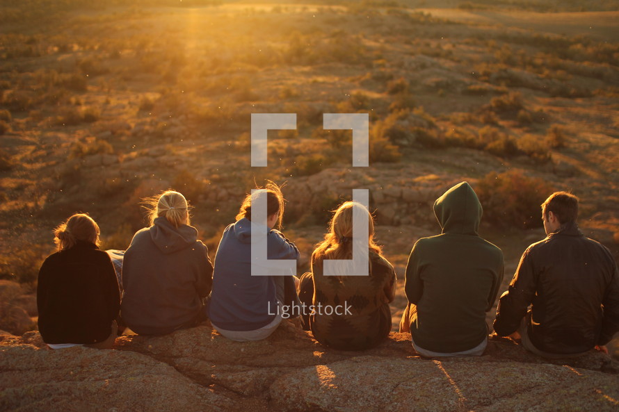 friends sitting at the edge of a cliff in a desert landscape at sunset 
