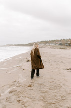 a woman walking in sand on a beach in a coat 