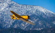 A yellow single engine airplane flying near a mountain.