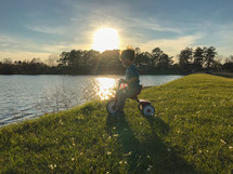 a toddler boy on a tricycle by a pond's edge 
