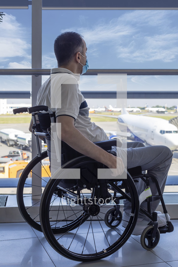 Young man with protective mask in a wheelchair at the airport looking out the window