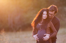 Expecting couple embracing outside at sunset.