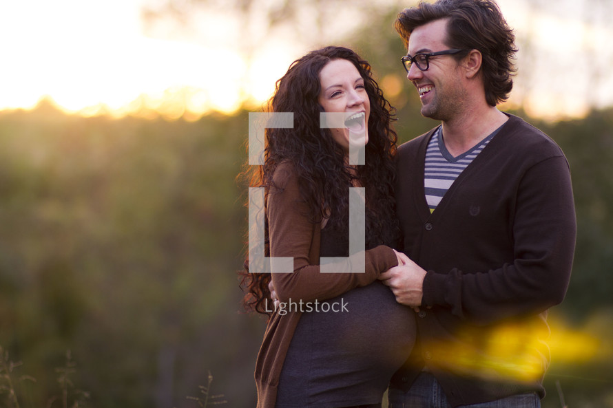 Expecting couple laughing while embracing outside at sunset.
