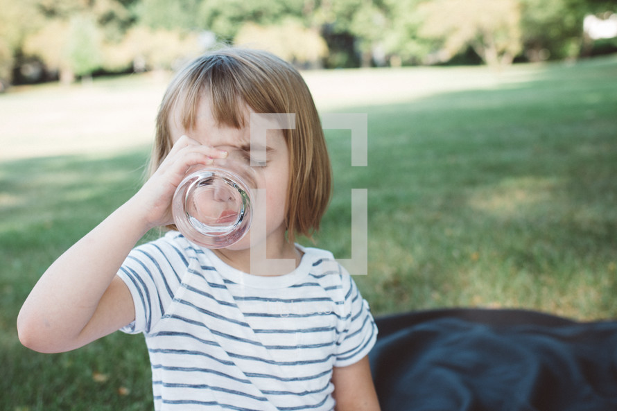 a girl child drinking out of a glass outdoors 