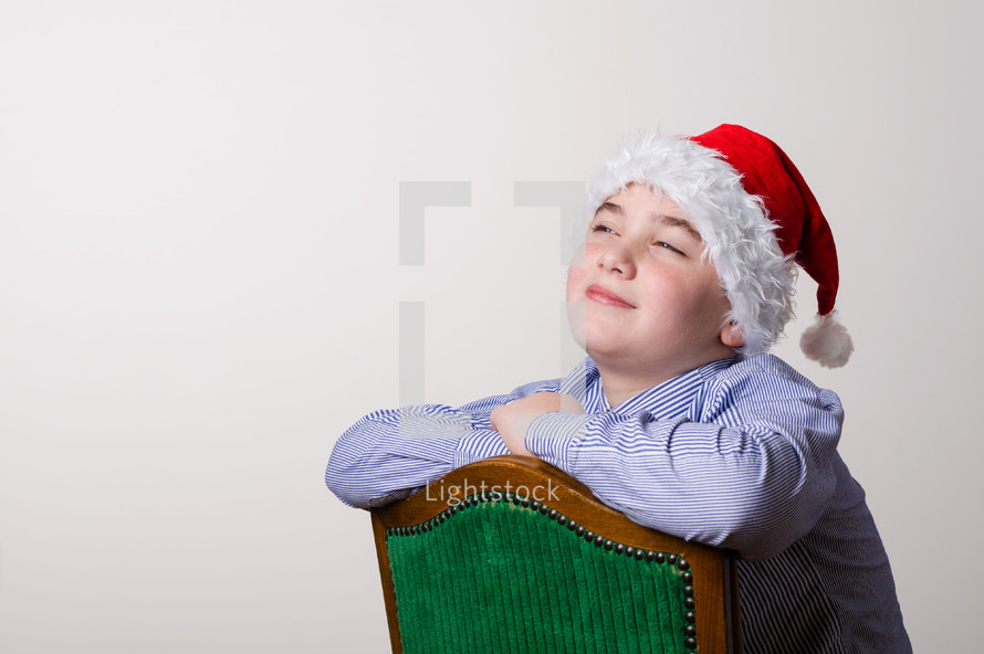 Boy wearing a Santa hat sitting in a chair, smiling pleasantly with his chin up.