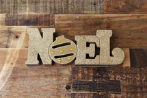 Word Noel in gold on a wooden floor type background 