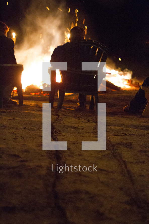 Man drags a Muskoka chair across a beach towards a large bonfire.