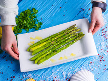 Female hands puts a plate of freshly prepared asparagus on a wooden blue table decorated with sea salt, herbs and spices. Beautiful Still Life, healthy lifestyle concept.